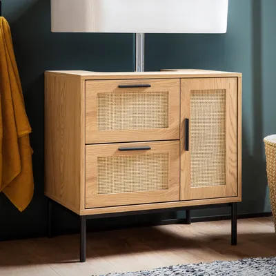 A wooden cabinet with rattan accents is shown in a bathroom setting, with a yellow towel hanging on the left and a basket on the right.