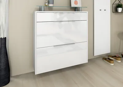 A white, high-gloss shoe cabinet with three drawers is mounted on a light beige wall. A black vase with white reeds is to the left, and a white panel with hooks is to the right. A few pairs of shoes are on the light wooden floor.