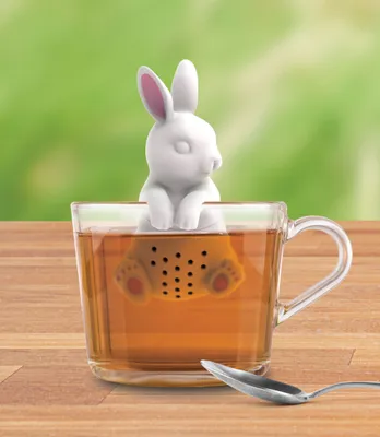 A white bunny-shaped tea infuser is sitting in a clear glass mug filled with tea, with a spoon on the side.