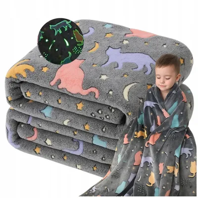 A child wrapped in a glow-in-the-dark cat-themed blanket, with a folded stack of the same blankets in the background.