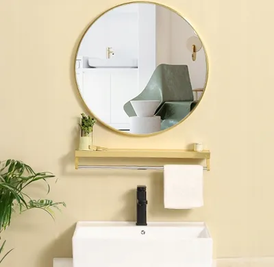A round mirror with a golden frame is mounted above a sink and shelf in a bathroom.