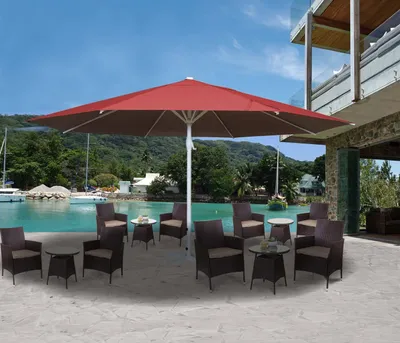 An outdoor patio scene with a large red umbrella, chairs, and tables set up near a body of water with boats and a building in the background.