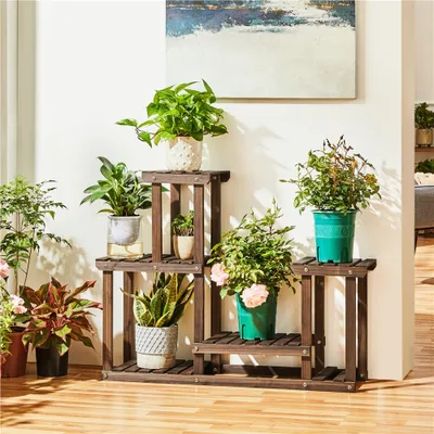 A wooden plant stand with multiple tiers, filled with various potted plants, is placed against a white wall with a painting above it.