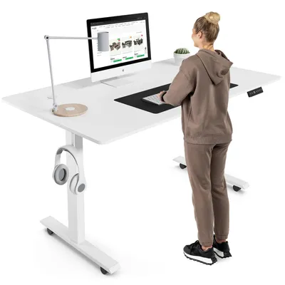 A woman stands at a white adjustable standing desk, using a computer. The desk has a lamp, a computer monitor, a keyboard, and headphones hanging from the side.