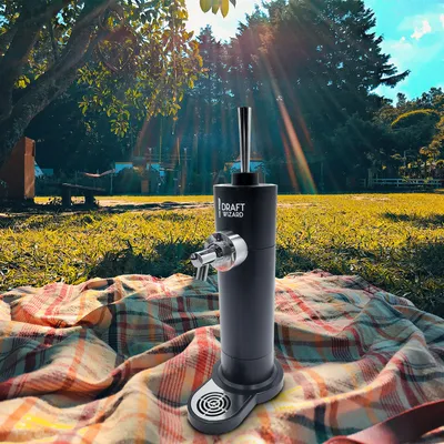 A beer dispenser is set up on a picnic blanket in a park setting with trees and sunlight.