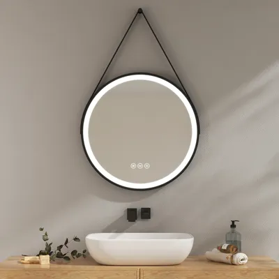 A round LED mirror with a black frame hangs above a white vessel sink on a wooden vanity. The mirror has a bright LED light around its edge and touch controls in the center. There are two black wall-mounted fixtures above the sink, and a soap dispenser and