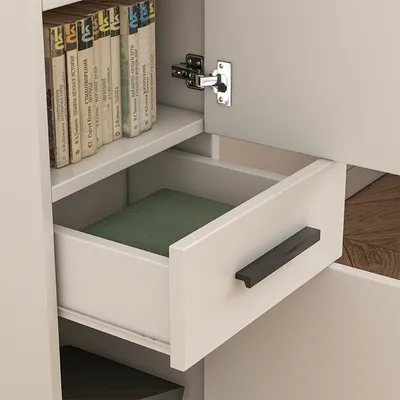 A close-up view of an open drawer in a white bookshelf, revealing a green book inside. A shelf above holds several books, and a cabinet hinge is visible on the right.