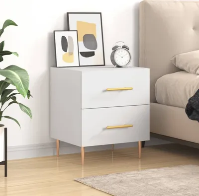 A white nightstand with gold handles is placed next to a bed. There are two framed pictures and an alarm clock on top of the nightstand. A plant is to the left of the nightstand.