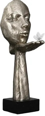 A silver-colored sculpture of a face with closed eyes, holding a delicate white butterfly in its open palm, mounted on a black base.