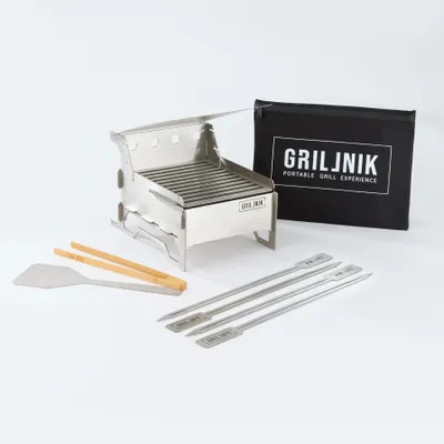 A portable stainless steel charcoal grill with accessories and a carrying bag, displayed on a white background.