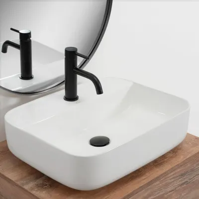 A white rectangular basin sink with a black faucet is shown on a wooden countertop, with a mirror reflecting the sink and faucet.
