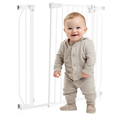A baby stands in front of a white baby gate.