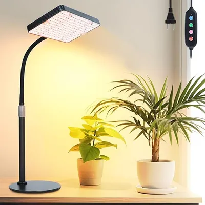 A grow light lamp is positioned on a desk next to two potted plants, with a power cord and controller visible in the background.