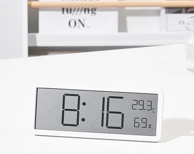 A digital clock displaying time, temperature, and humidity on a white desk.