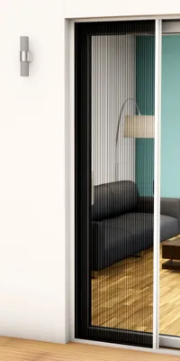 A close-up view of a white pleated door screen with a black frame, set against a white wall and a glimpse of a living room interior.
