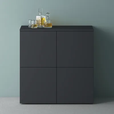 A dark gray cabinet with four doors is shown against a muted blue-green wall and a gray floor. On top of the cabinet are a decanter and three glasses filled with a golden liquid, suggesting a bar or storage area.