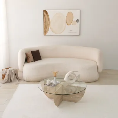 A modern living room scene with a fluffy cream couch, a glass coffee table with a travertine base, and abstract art on the wall.