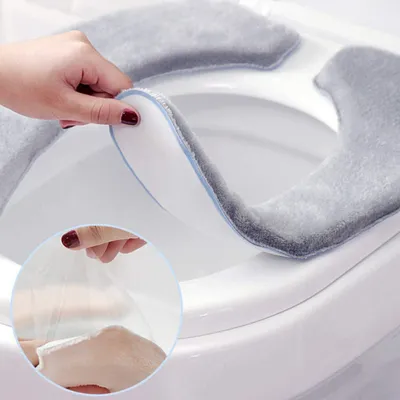 A hand is shown attaching a soft, gray toilet seat cover to a white toilet seat. A close-up inset shows the adhesive backing of the cover being peeled off.