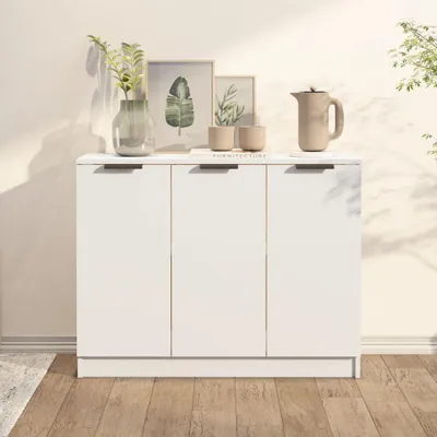 A white sideboard with three doors is shown against a light-colored wall. The top of the sideboard is decorated with a vase of plants, two small cups, and a pitcher. The floor is made of wooden planks.