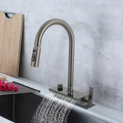 A modern kitchen faucet with a pull-down sprayer and waterfall spout is shown in use, with water flowing into a sink. A cutting board and grapes are visible in the background.