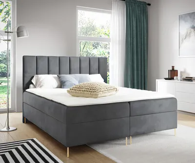 A bedroom scene featuring a large gray upholstered bed with gold legs, set against a window with green curtains.