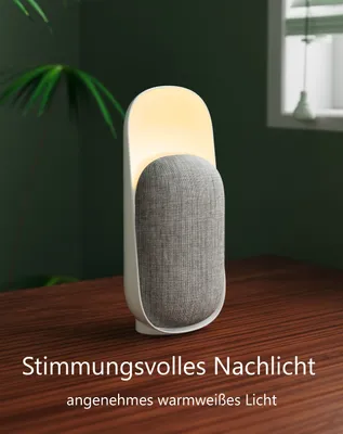 A bedside lamp with a fabric speaker sits on a wooden table, with a plant and window in the background.