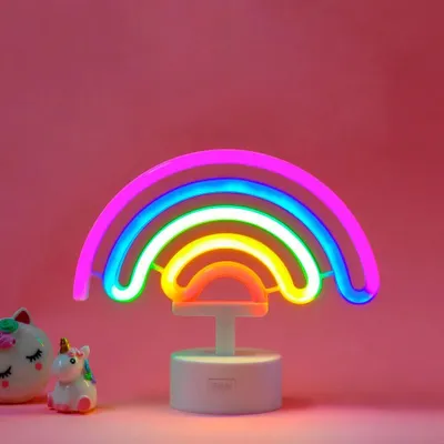 A neon rainbow lamp with a white base, placed on a pink background. Two small decorative unicorn figures are visible to the left of the lamp.