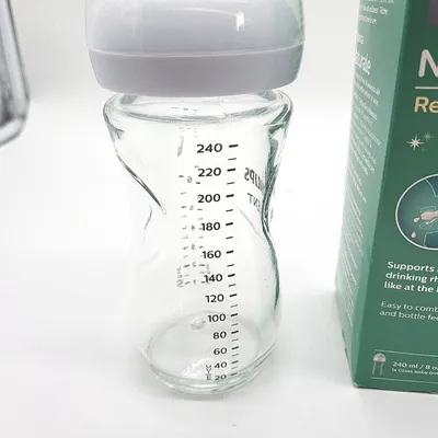 A close-up of a glass baby bottle with measurement markings and a white cap, next to a green product box.