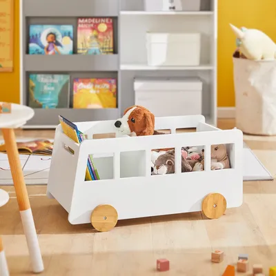 A white toy bus storage unit filled with stuffed animals and books, placed in a child's room with a bookshelf in the background.