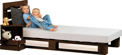 Two children are sitting on a wooden pallet bed with a white mattress.