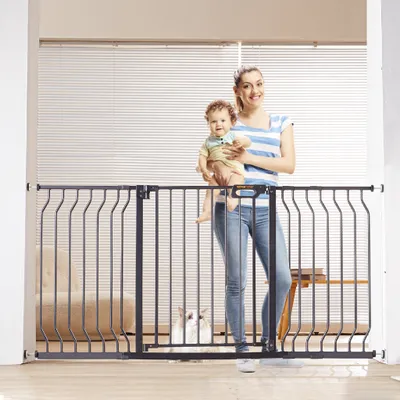 A woman holds a baby while standing behind a black metal baby gate. A cat is visible behind the gate.