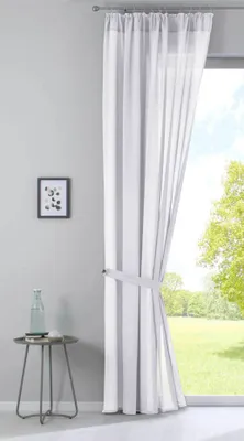 A sheer white curtain with gray stripes is tied back with a fabric band, hanging in front of a window with a view of a green lawn and trees. A small side table with a bottle and a framed picture on the wall are also visible.
