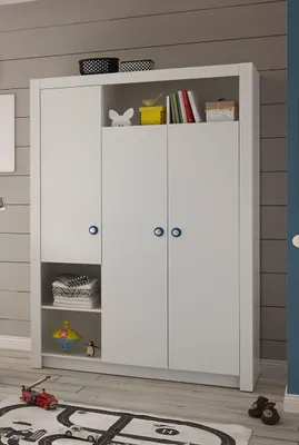 A white three-door wardrobe with shelves and decorative items on top, placed in a room with gray paneled walls and a rug on the floor.