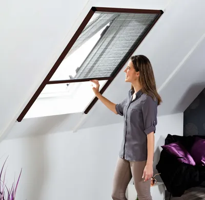 A woman is adjusting a screen on a skylight window in an attic room.