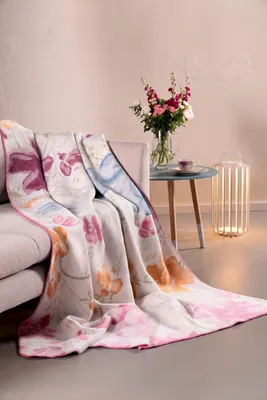 A floral blanket draped over a sofa next to a side table with a vase of flowers and a lamp.