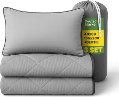A folded bedding set including a pillow and two folded blankets, neatly packed in a gray drawstring bag. A tag with product details is attached to the bag.