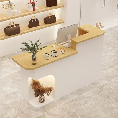 A retail store counter with handbags displayed on shelves behind it. The counter has a computer monitor, payment devices, and a vase of greenery.