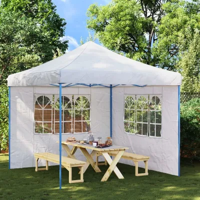A white party tent with windows is set up in a garden, with a wooden picnic table and benches inside.