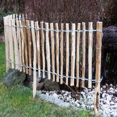 A rustic wooden fence made of natural hazelnut branches, bordering a garden area with grass, rocks, and plants.