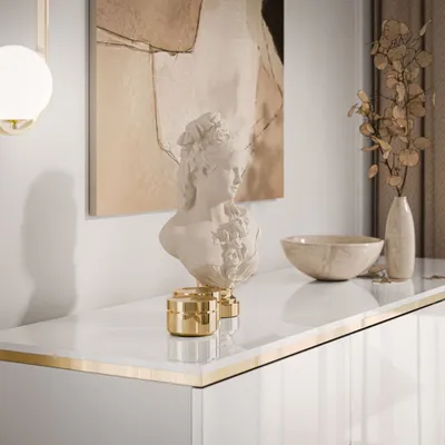 A white glossy dresser with gold trim, decorated with a bust, a bowl, and dried flowers in a vase.