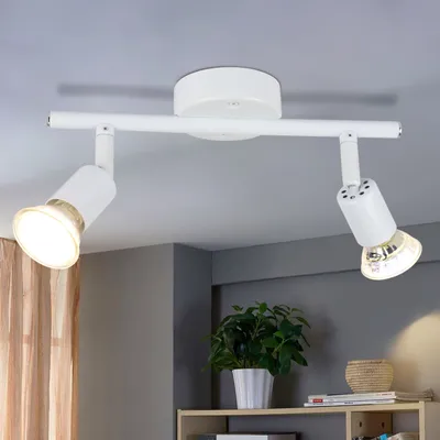 A white LED ceiling spotlight with two adjustable heads is mounted on a white track attached to a white ceiling. The background includes a gray wall and a sheer brown curtain.