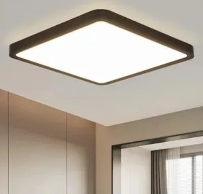 A square LED ceiling light fixture mounted on a ceiling, with a dark frame and a bright white light panel. The background shows a hint of a wall and a doorway.