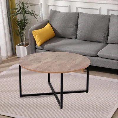 A round coffee table with a wood-look top and black metal legs is placed on a rug in front of a gray sofa. A yellow pillow is on the sofa, and a potted plant is to the left.