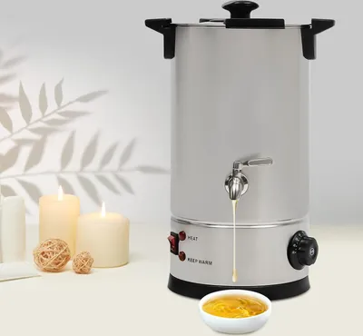 A silver wax melting pot with a spigot is shown on a table with candles and decorative balls in the background.