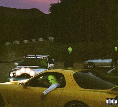 Several people wearing green ski masks are standing around and in cars at dusk.