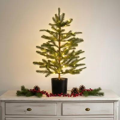 A small, decorated artificial Christmas tree with LED lights sits on a white dresser, adorned with a garland of pine sprigs, berries, and pinecones.