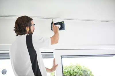 A man is using a cordless drill to attach something to a ceiling track.