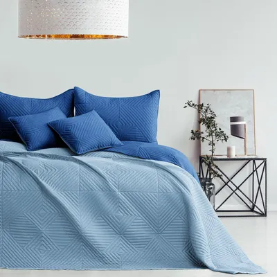 A bed with blue bedding and pillows, in a room with a side table and lamp.