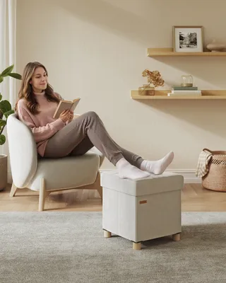 A woman is sitting in a chair reading a book, with her feet resting on an ottoman. The scene is set in a living room with shelves on the wall.