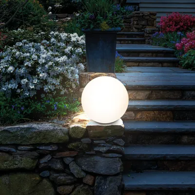 A glowing spherical solar light sits on a stone wall next to a garden path with flowering bushes.